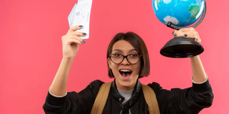 Jovem estudante ergue passagens aéreas e um mapa em formato de globo com as mãos enquanto faz expressão de euforia. Falando sobre bolsas de estudo para brasileiros.