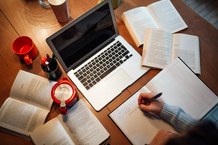 Mesa de estudos repleta de materiais, uma xícara e um notebook. Há também a mão de um estudante fazendo anotações. Falando sobre como passar em um concurso público.