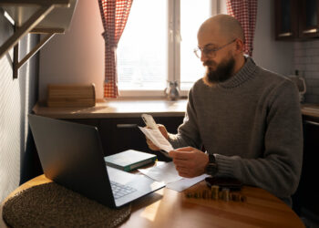 Homem sentado em mesa com laptop e documentos, analisando finanças para MEI.