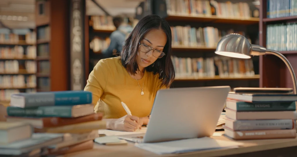 Mulher estuda para concursos com a ajuda de livros e um notebook. Falando sobre materiais gratuitos para concursos.