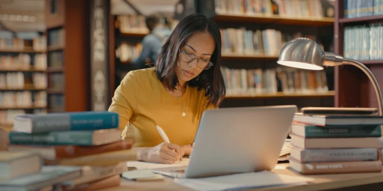 Mulher estuda para concursos com a ajuda de livros e um notebook. Falando sobre materiais gratuitos para concursos.