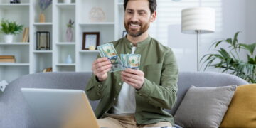 Os 7 melhores tipos de empréstimo para resolver emergências em 2025 3 Homem sorridente conta cédulas enquanto está sentado em um sofá em frente a um notebook. Falando sobre os melhores tipos de empréstimo.