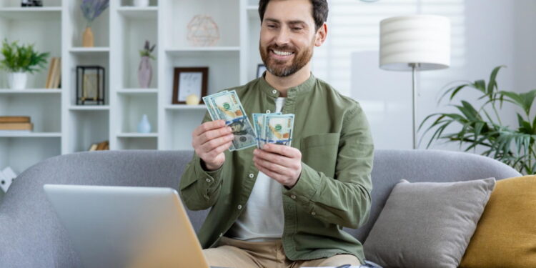 Os 7 melhores tipos de empréstimo para resolver emergências em 2025 16 Homem sorridente conta cédulas enquanto está sentado em um sofá em frente a um notebook. Falando sobre os melhores tipos de empréstimo.