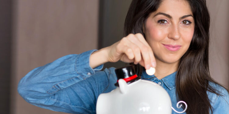 Método 50-30-20: Veja Como Usar para Manter seu Orçamento Sob Controle 18 Uma mulher sorridente colocando moedas em um cofrinho, ilustrando o método 50-30-20 de finanças pessoais.